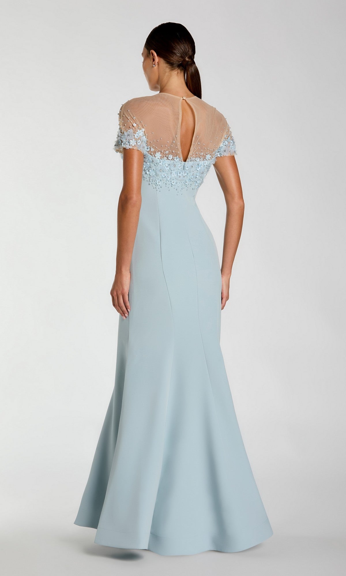 A woman faces away, wearing the Mac Duggal 11867 Short Sleeve Long Formal Dress, a floor-length, light blue beaded gown with a sheer embellished bodice and keyhole back detail.